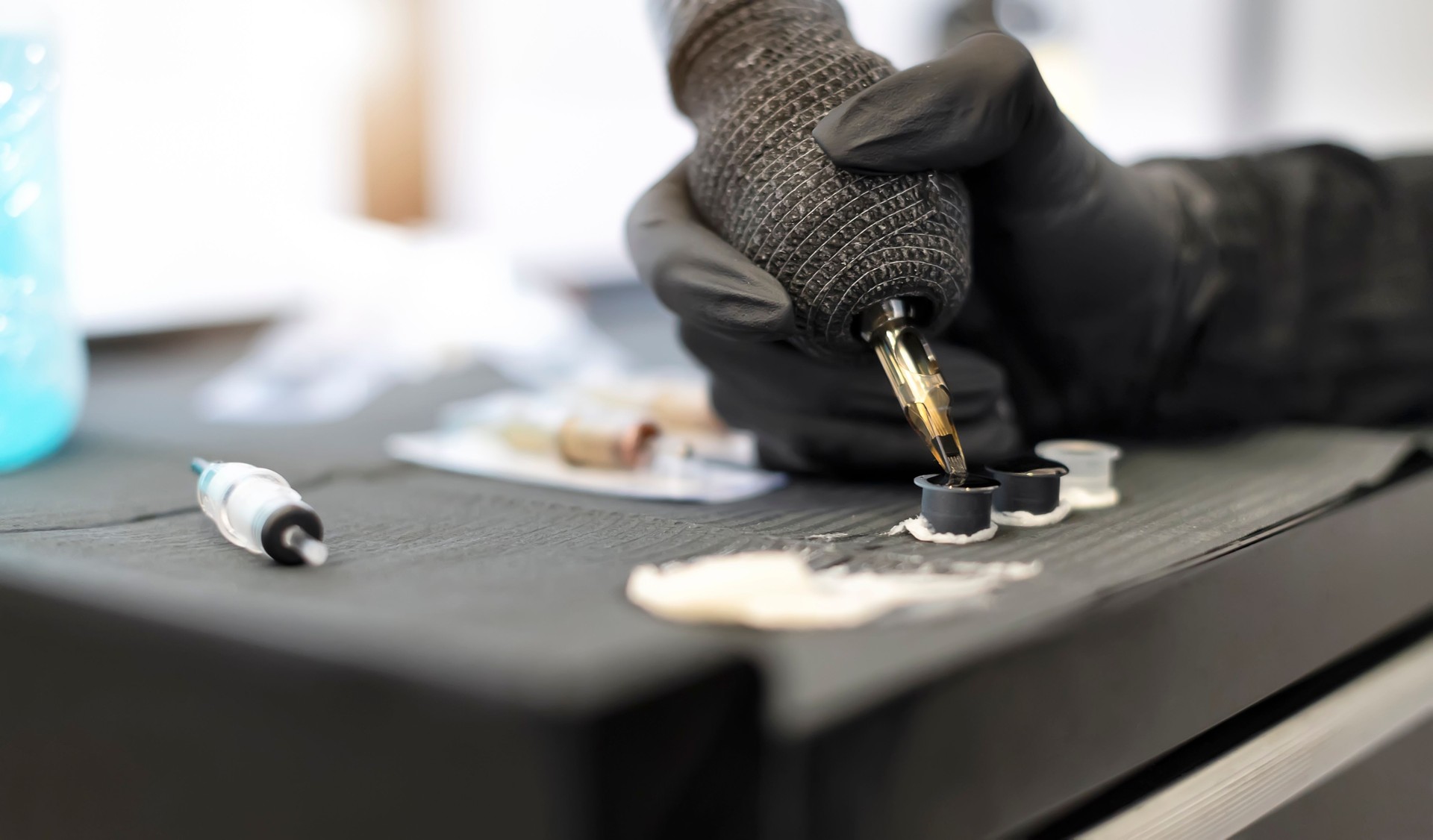 Close-up professional female tattoo artist in a black glove with a tattoo machine picks up black ink from a jar - Concept about equipment for making tattoo art